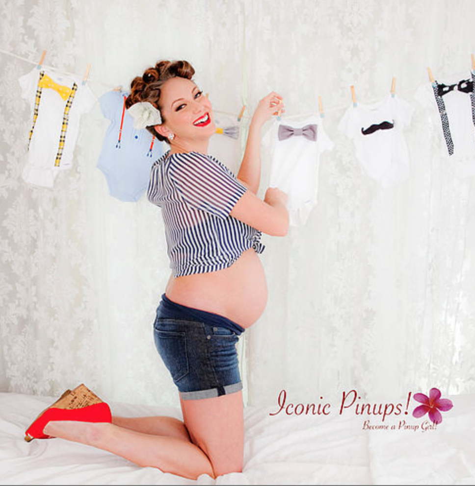 Photo of cute Rosie the Riveter style pinup girl doing a mom to be maternity photo shoot , complete with baby clothes next to a lace curtain
