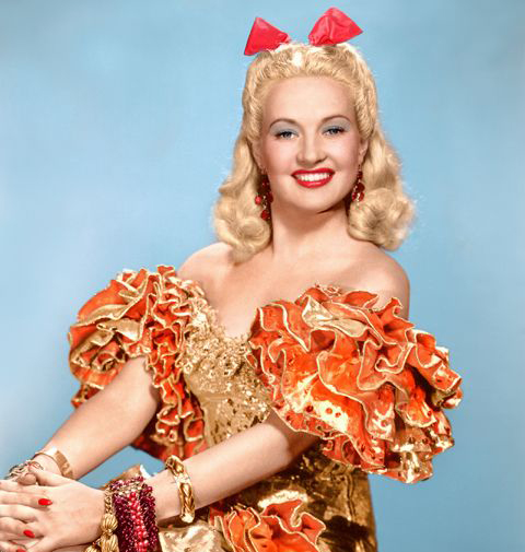 technicolor photo of Betty Grable. She has an orange and gold cuban style costume with ruffled sleeves, a hair bow, and vintage red lipstick.
